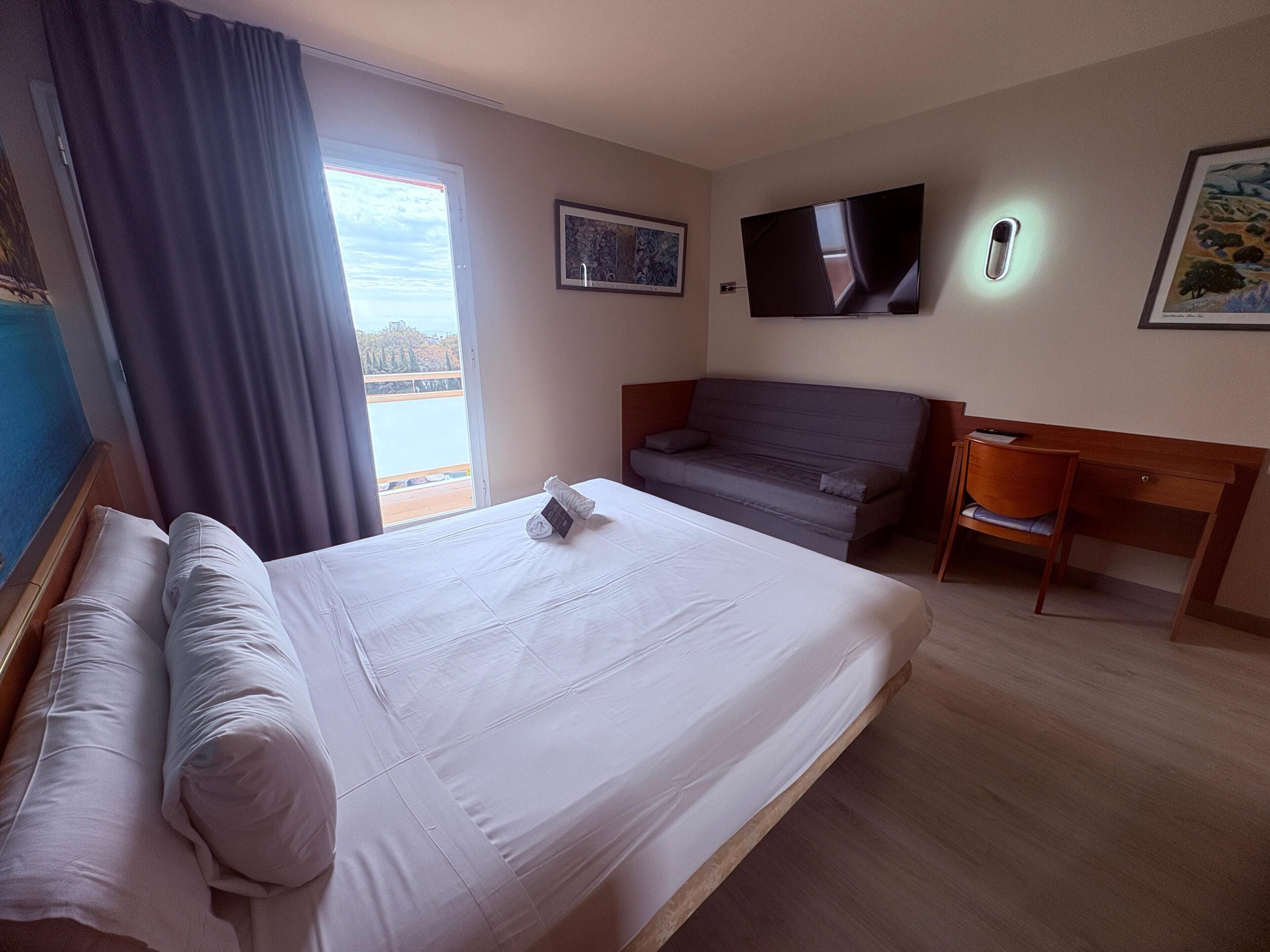 A hotel room with a bed, a sofa, a desk, a TV, and a window with a view.