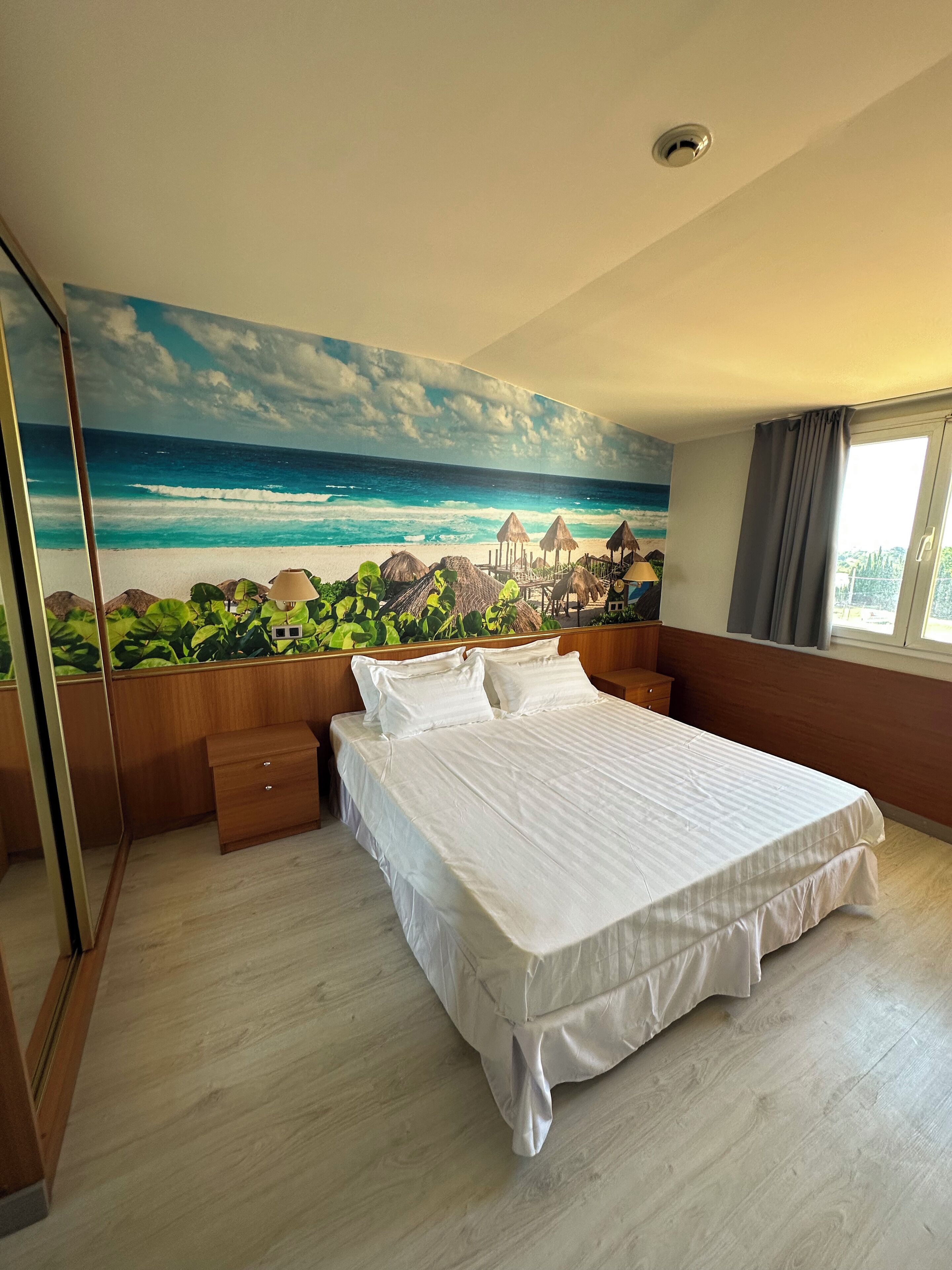Un dormitorio con cama, mesitas de noche de madera, un ventanal con cortinas y un mural de playa en la pared.