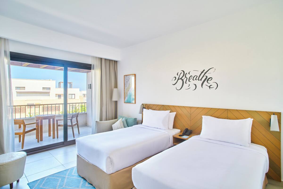 Une chambre d’hôtel avec deux lits, un balcon offrant une vue et un mur sur lequel est écrit le mot « Respirez ».