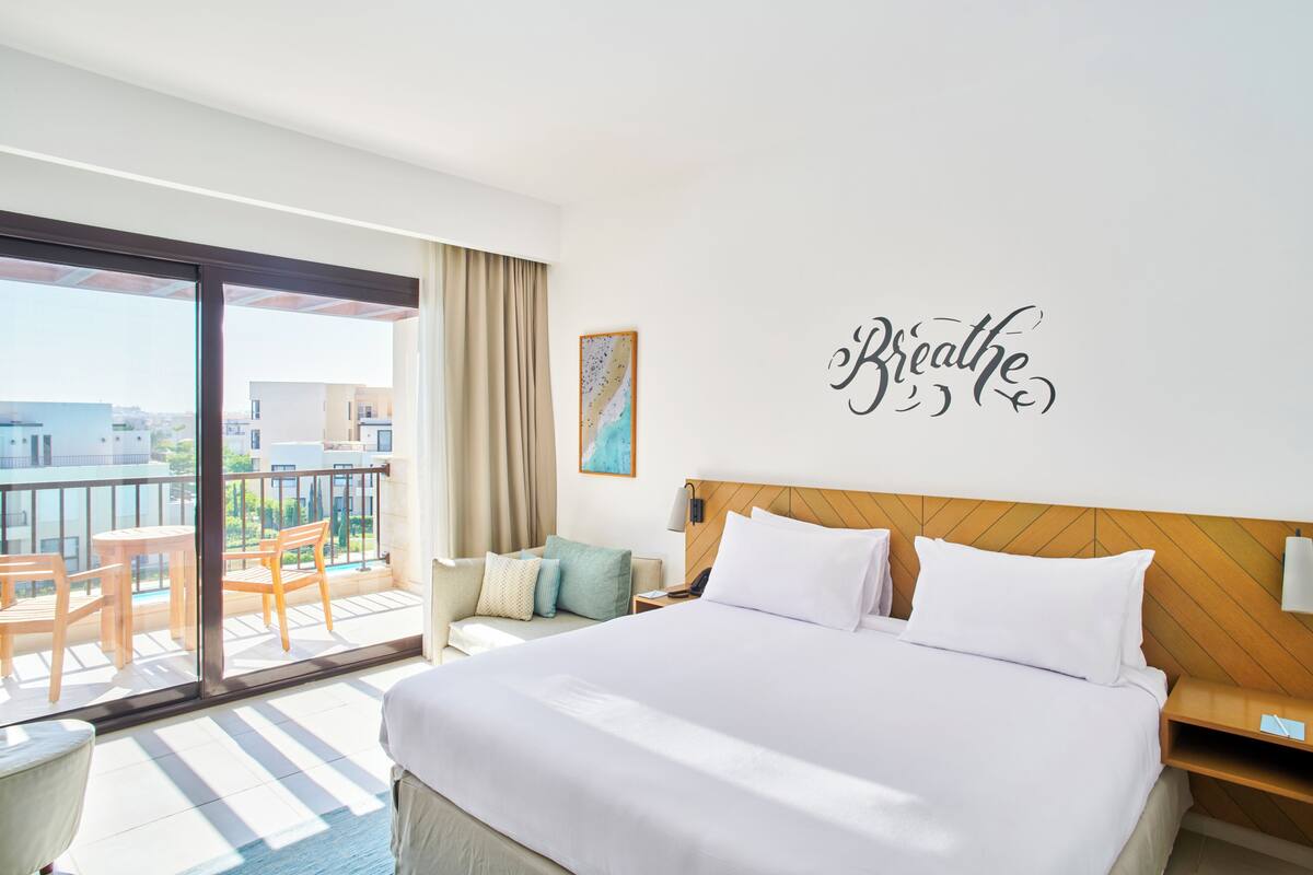 Une chambre d’hôtel avec un grand lit, un balcon avec des chaises et un mur sur lequel est écrit le mot « Respirez ».