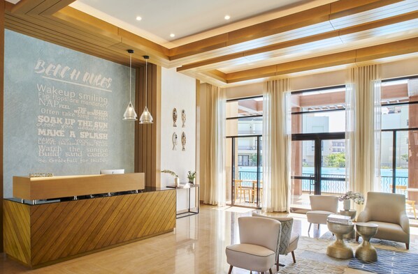 A hotel reception area with a wooden reception desk, a chalkboard with beach rules, and a view of the pool through large windows. - Marassi Boutique Hotel-Blanca (El Alamein)