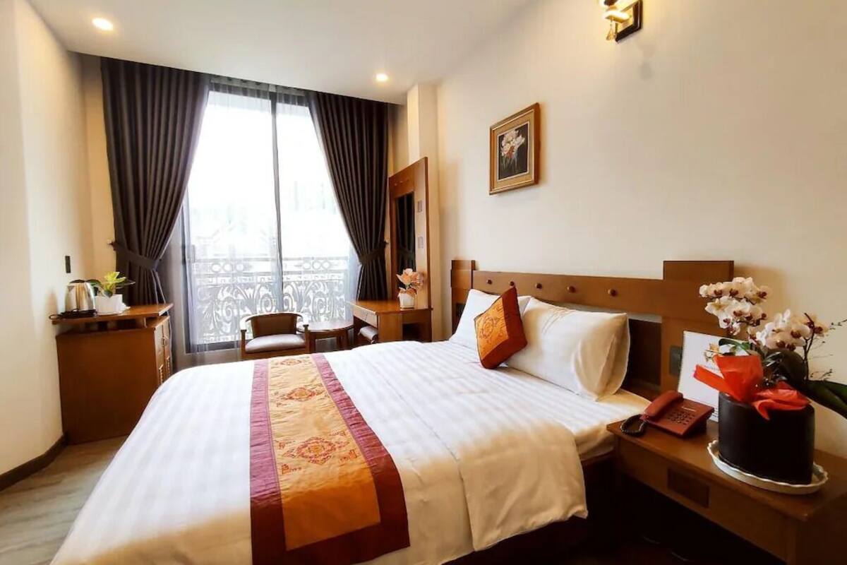 A hotel room with a large bed, a desk with a telephone, a chair, a window with curtains, and a wall-mounted light fixture.
