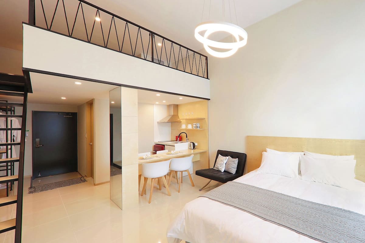 Ein modernes Hotelzimmer mit einem großen Bett, einem Schreibtisch mit zwei Stühlen, einer Küchenzeile und einem Balkon mit Geländer.