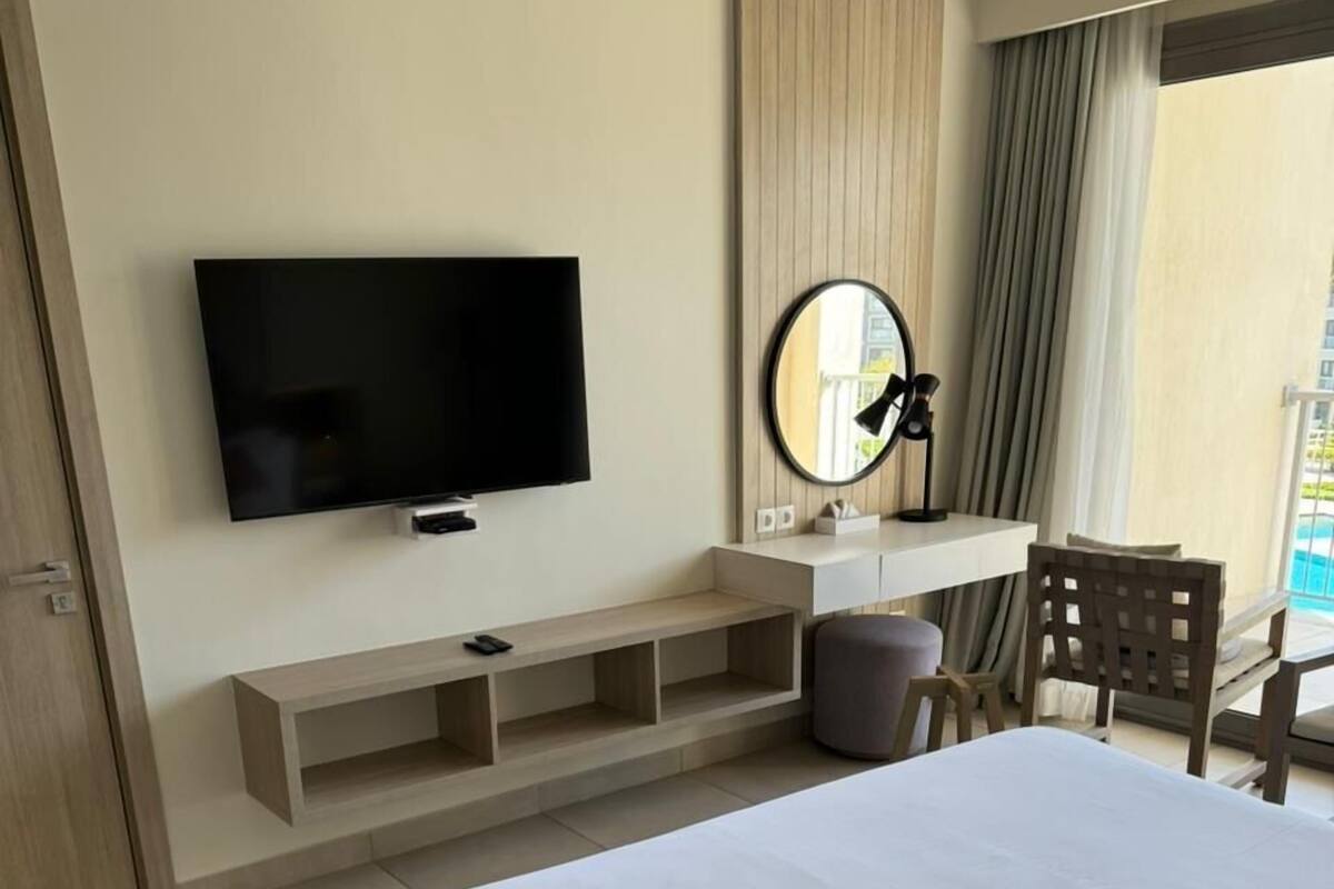 Een hotelkamer met een groot bed, een aan de muur bevestigde televisie, een bureau met stoel en een raam met gordijnen.