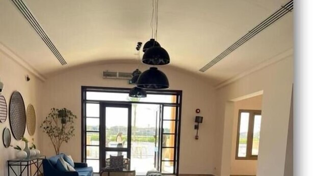 Ein geräumiges Wohnzimmer mit großem Fenster, einem blauen Sofa, gemusterten Hocker und einer Pendelleuchte.
