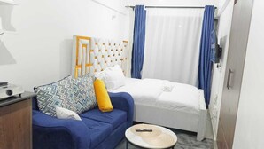A hotel room with a bed, a blue sofa with patterned cushions, a small round table, and a television. - Lux Suites Tsavo Sunset Apartments  (Nairobi)