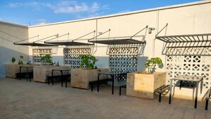 An outdoor patio area with multiple tables and chairs, some with attached umbrellas. There are potted plants and a decorative white lattice structure. - Lux Suites Tsavo Sunset Apartments  (Nairobi)