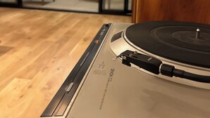 Un tocadiscos en una habitación con suelo de madera, un ventanal corredero que da a un balcón con una silla y una mesa.