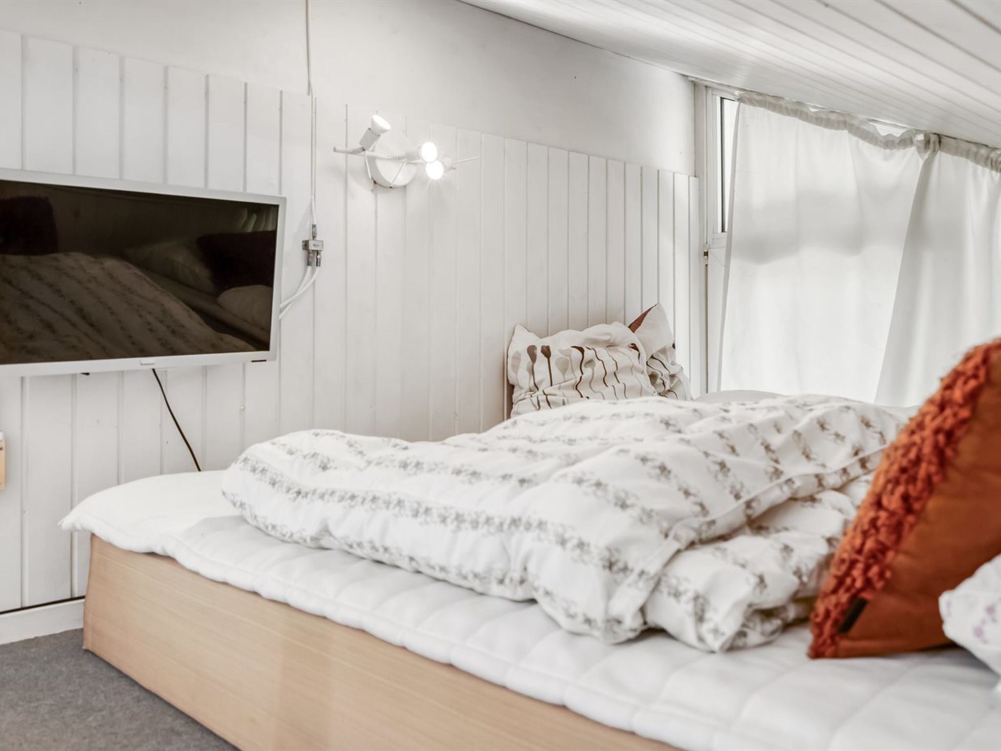 A bedroom with a bed, a mounted TV, and a window with curtains.