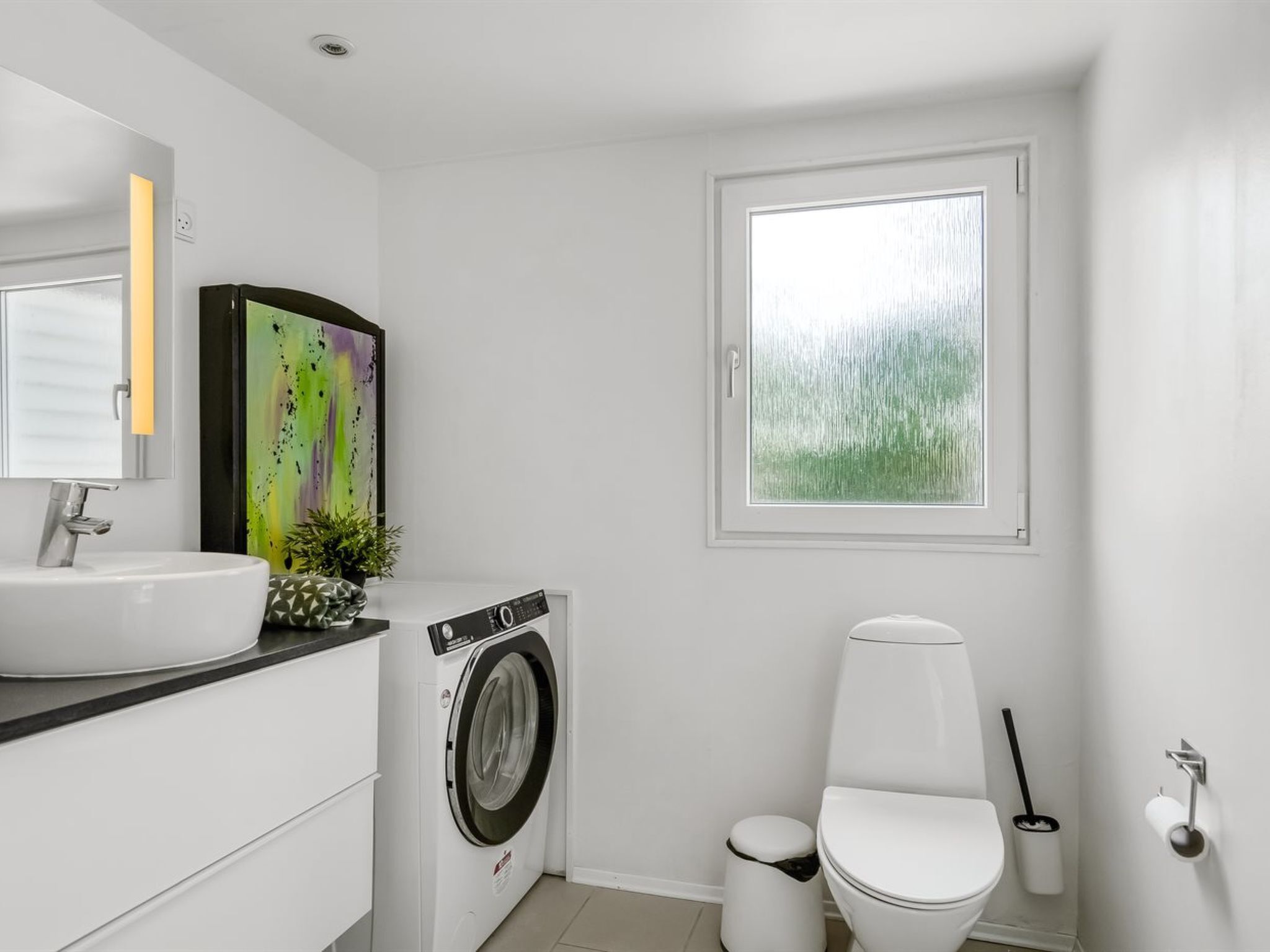 Une salle de bain moderne avec un lavabo blanc, un lave-linge, des toilettes et une fenêtre donnant sur un espace verdoyant.