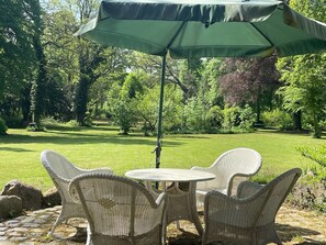 A patio with wicker chairs and a table under a green umbrella, surrounded by a lush garden.
