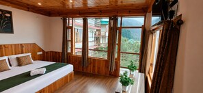 A room with a bed, wooden walls, large windows, and a view of a building and mountains. - Mahamaya Resort (Manali)