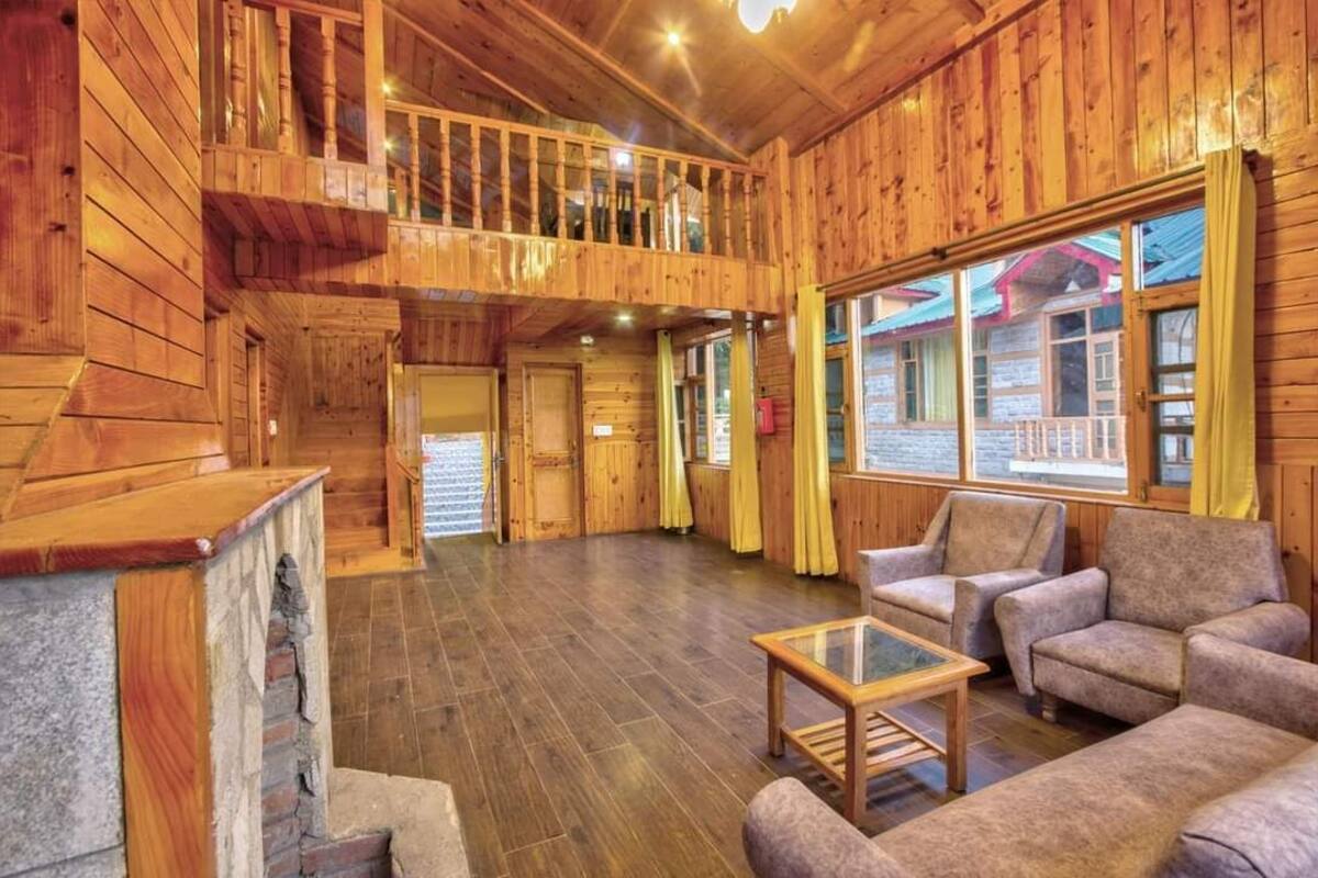 A wooden cabin interior with a balcony, a stone fireplace, and a large window with curtains.