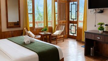 A hotel room with a large bed, wooden flooring, a desk, a chair, a television, and a window with curtains.