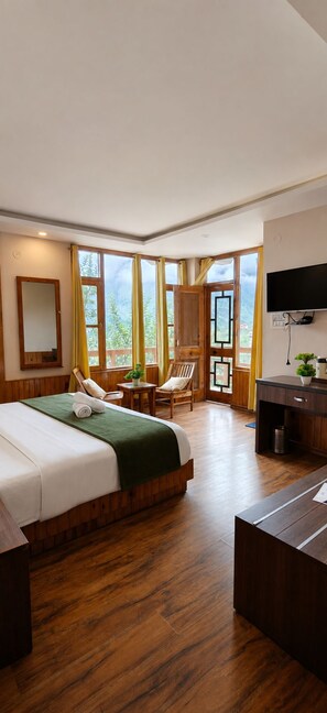 A hotel room with a large bed, wooden flooring, a desk, a chair, a television, and a window with curtains. - Mahamaya Resort (Manali)