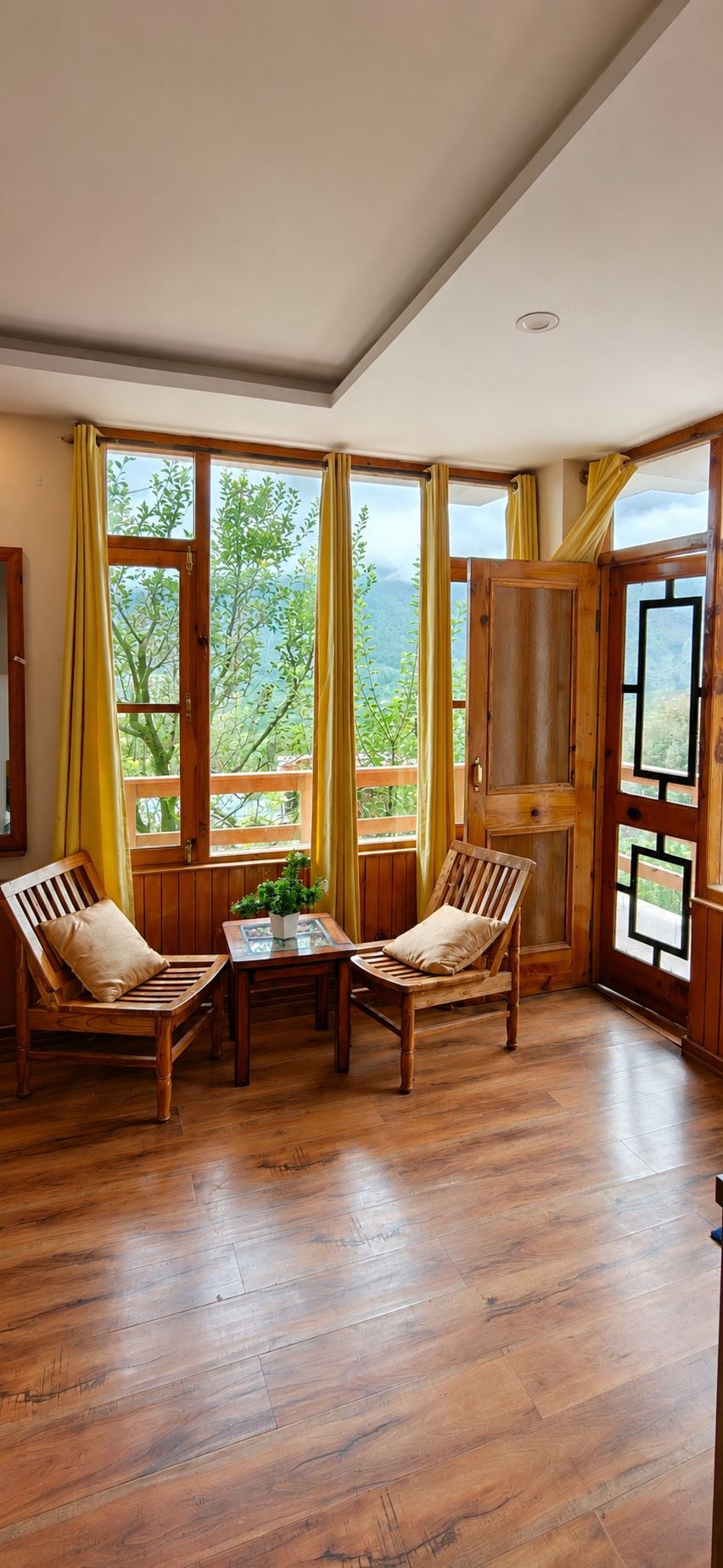 Una habitación con piso de madera, dos sillas de madera, una pequeña mesa con una planta y ventanas grandes con cortinas.