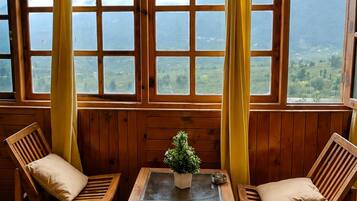 Ein Zimmer mit zwei Holzsesseln, einem kleinen Tisch und Blick auf eine gebirgige Landschaft durch große Fenster mit gelben Vorhängen.