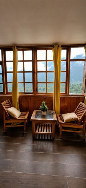 A room with two wooden chairs, a small table, and a view of a mountainous landscape through large windows with yellow curtains. - Mahamaya Resort (Manali)