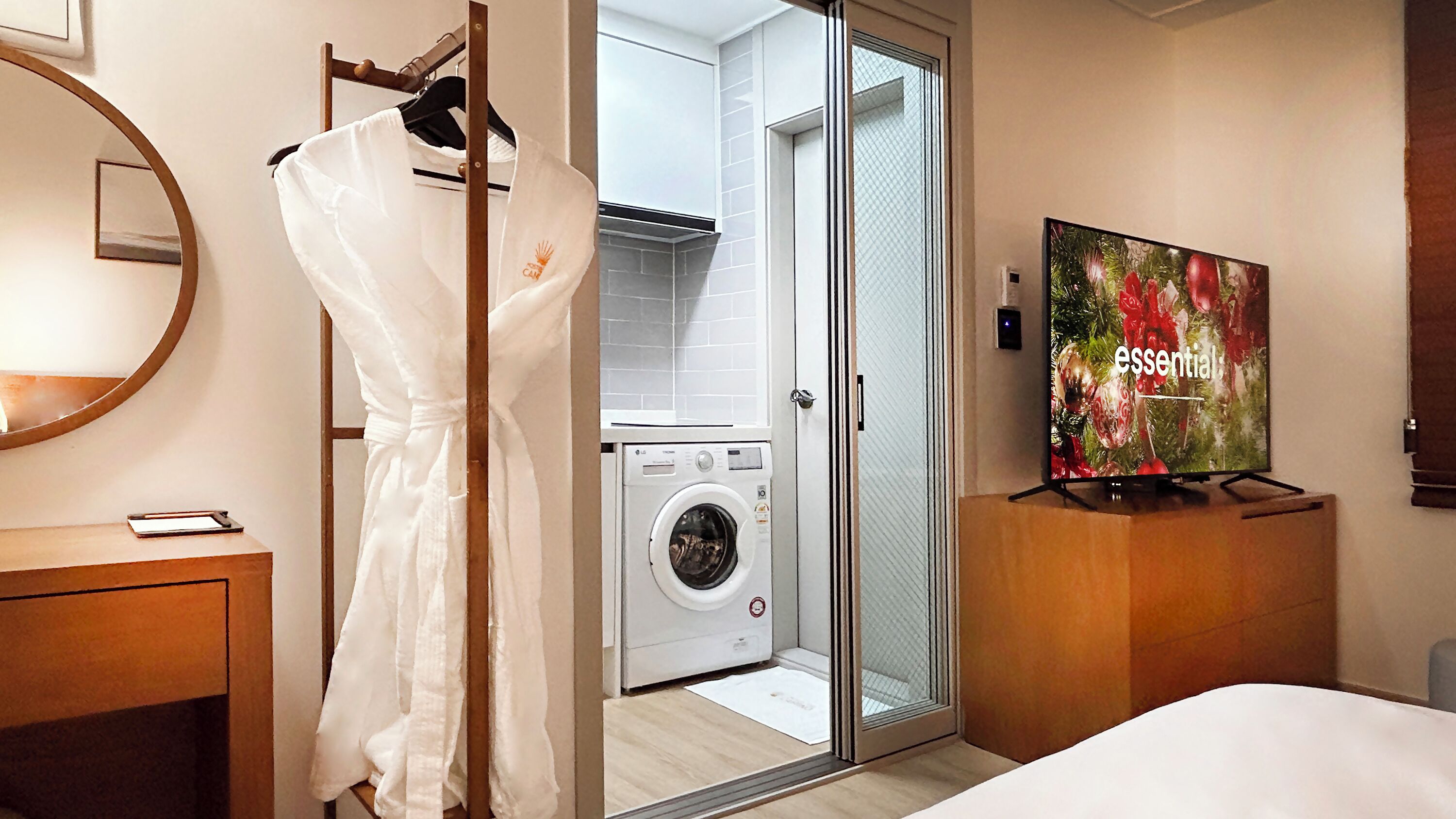 A hotel room with a washing machine, a robe hanging on a rack, a bed, a television, and a mirror.