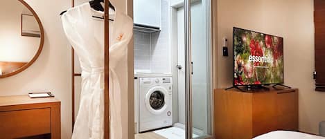 A hotel room with a washing machine, a robe hanging on a rack, a bed, a television, and a mirror.