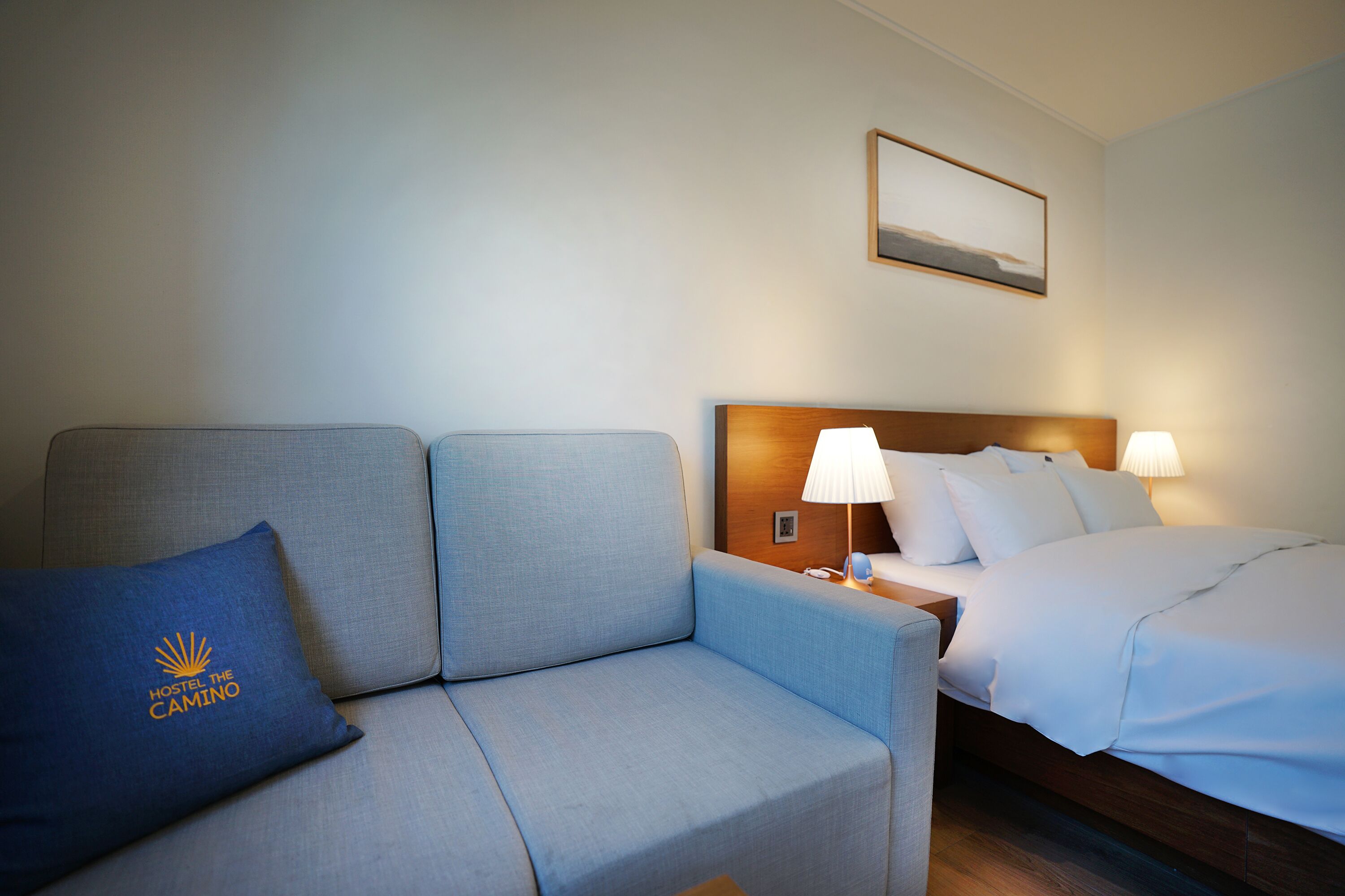 A hotel room with a grey sofa, a bed with white bedding, a nightstand, a lamp, and a wall-mounted picture.