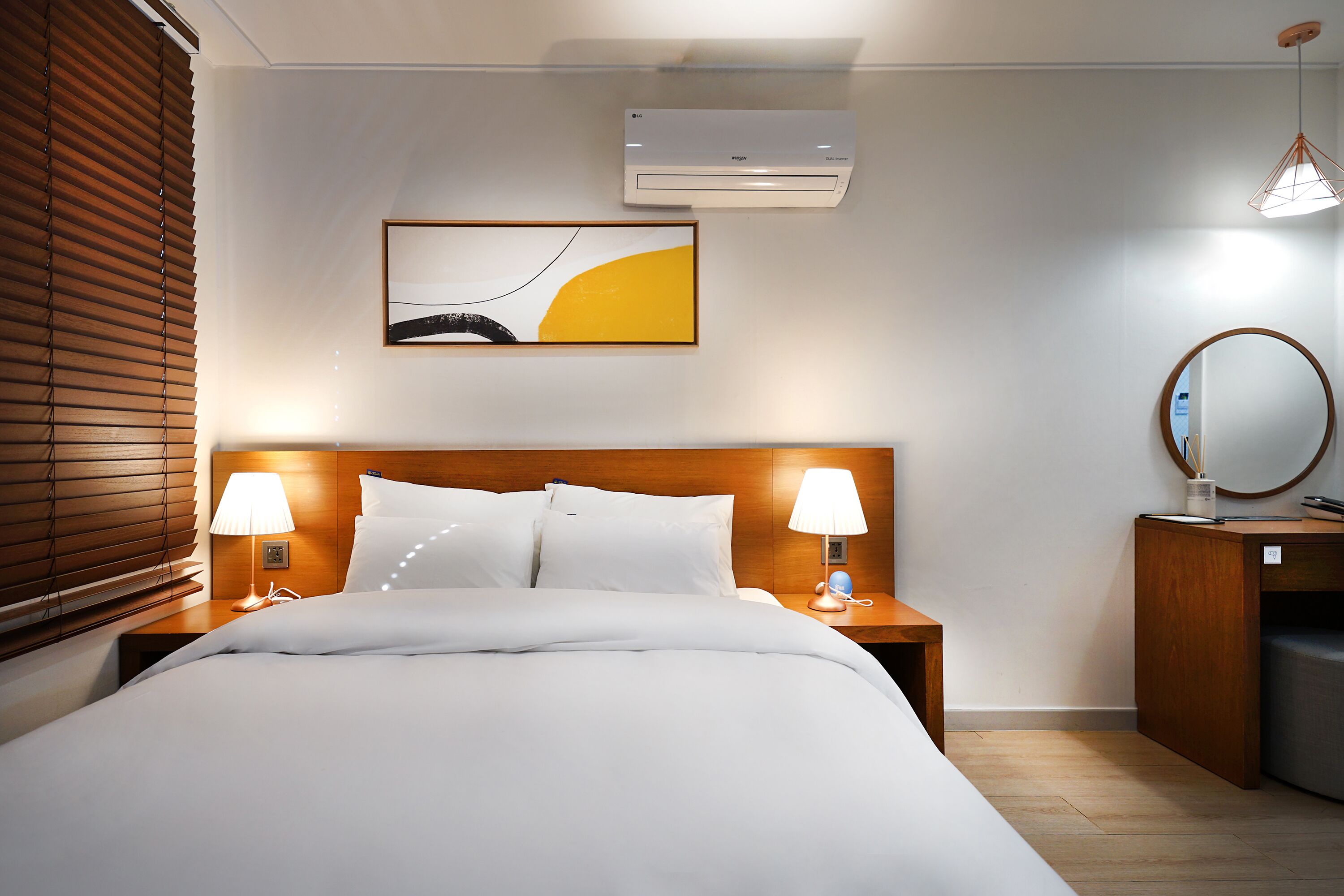 A hotel room with a bed, two bedside lamps, a wall-mounted air conditioner, a painting, a mirror, and a wooden nightstand.