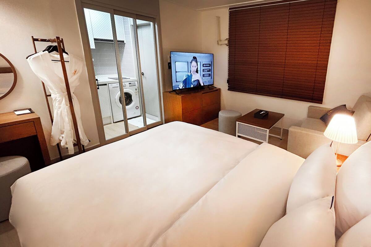 Une chambre d’hôtel avec un grand lit, une télévision, un bureau avec une lampe, une chaise et une fenêtre avec des stores.