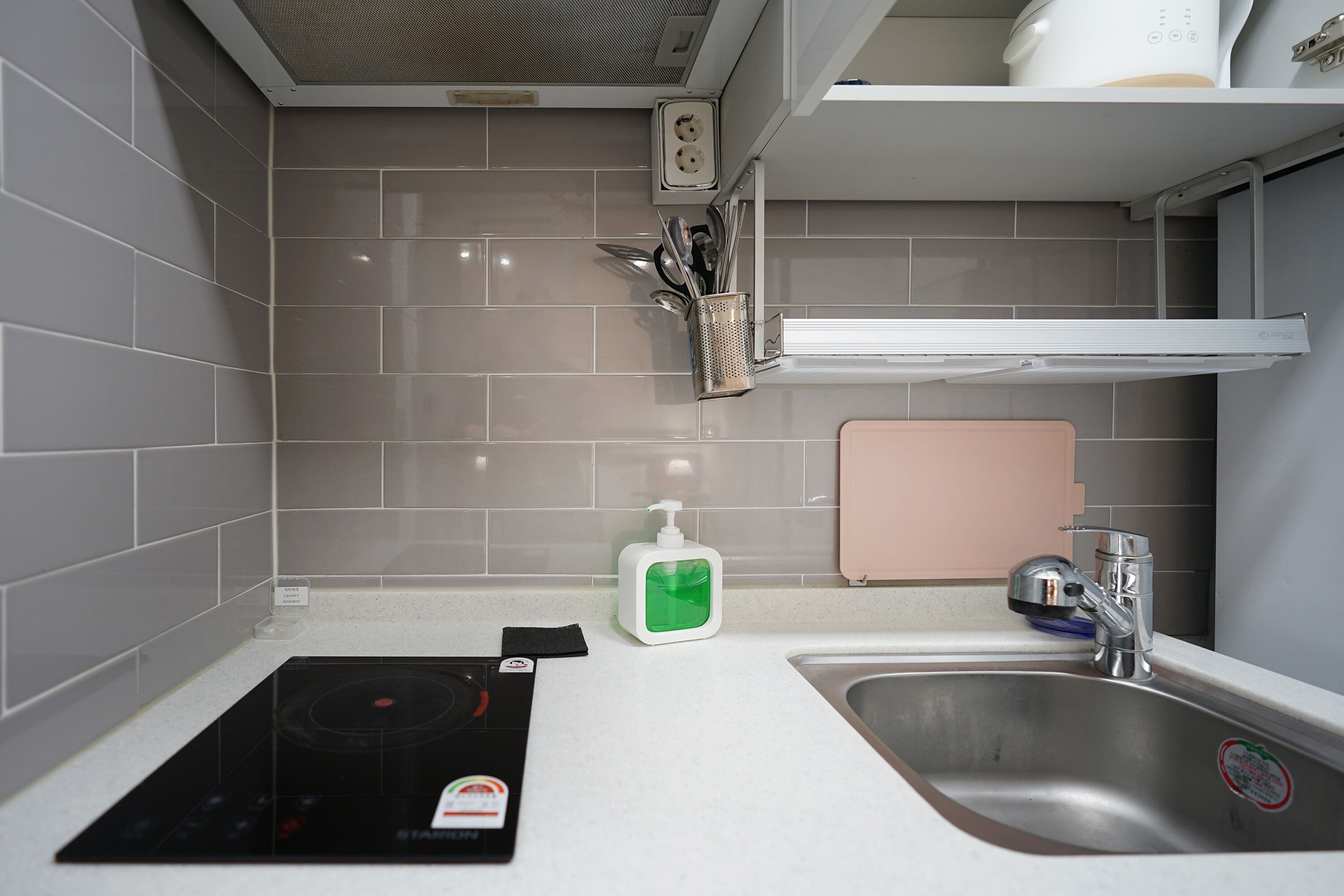 Een moderne keuken met grijze tegels, een ingebouwde kookplaat, een spoelbak en een plank met keukengerei.