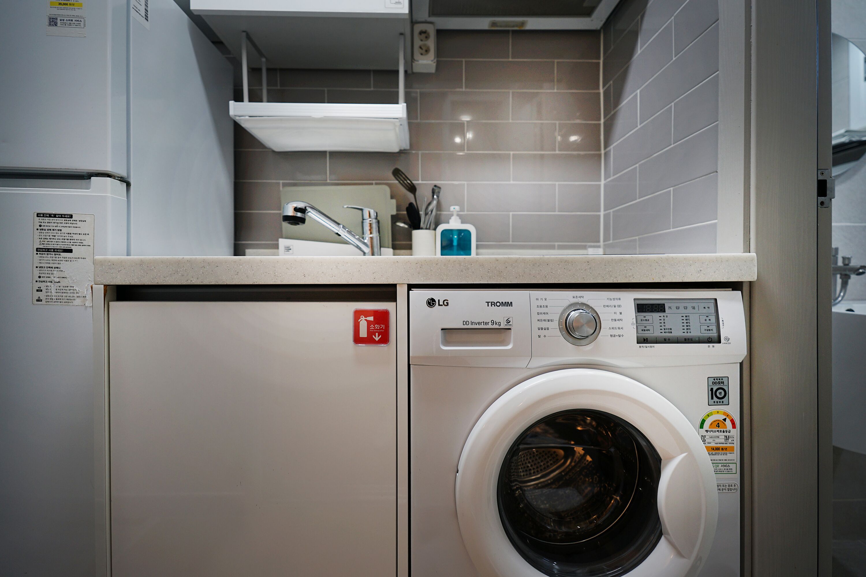 A compact laundry room with a front-loading washing machine, a small shelf above the machine, and a fire extinguisher sign.