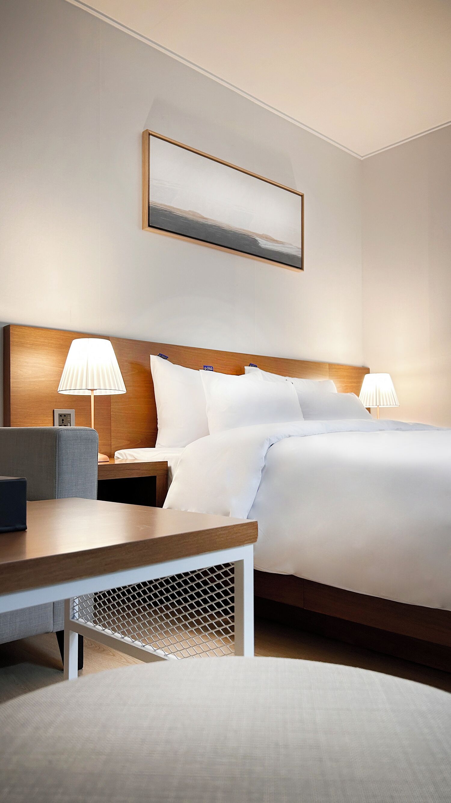 A hotel room with a large bed, two bedside tables with lamps, a wooden headboard, a small desk, and a wall-mounted picture.