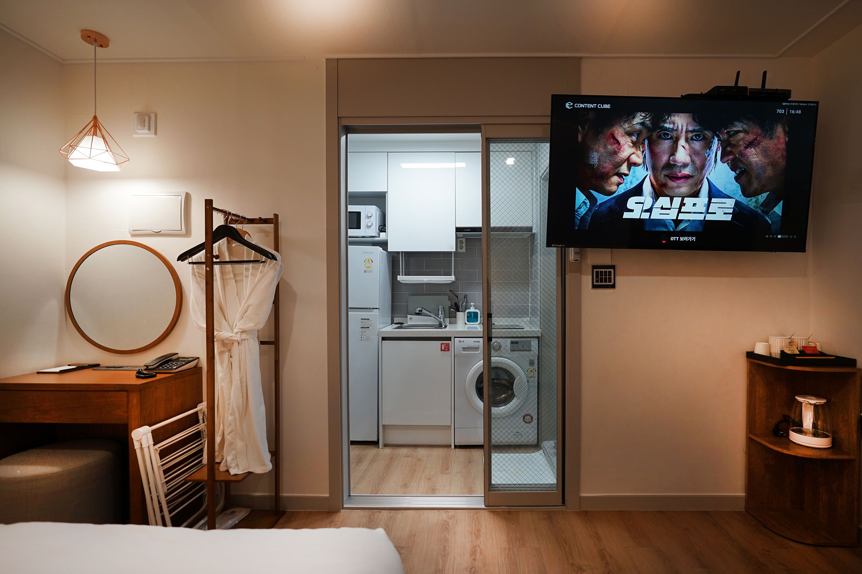 Ein Hotelzimmer mit Fernseher, der einen Film zeigt, einem Kleiderschrank, einem Spiegel, einem Schreibtisch mit Telefon und einer Waschmaschine.