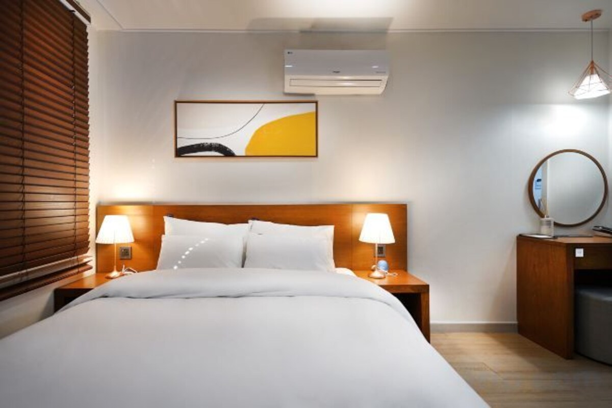 Een hotelkamer met een bed, twee bedlampen, een wandairconditioner, een schilderij, een spiegel en een nachtkastje van hout.