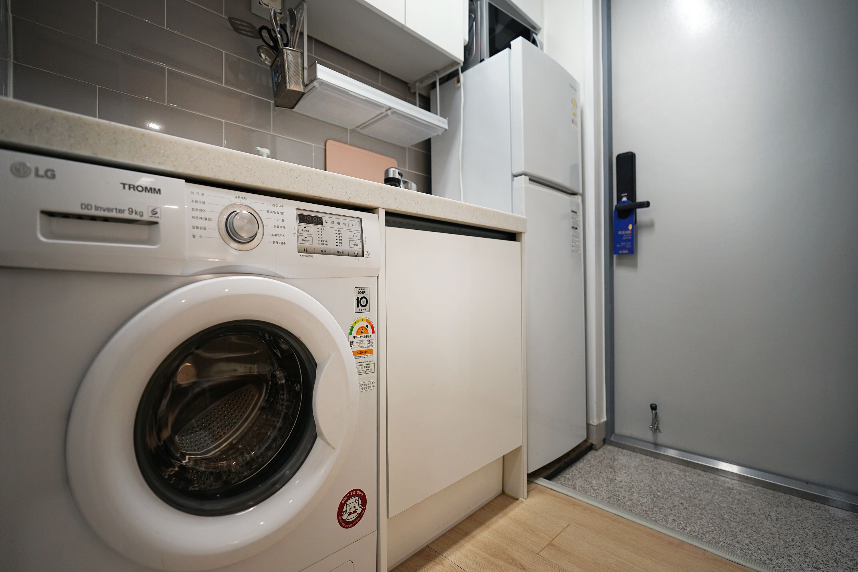 Un lavadero pequeño con una lavadora LG TROMM de carga frontal, un refrigerador compacto y una puerta con cerradura azul.