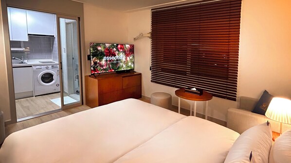 A hotel room with a bed, a TV, a small table, a lamp, and a view of a laundry area. - Hostel The Camino Seoul (Seoul)