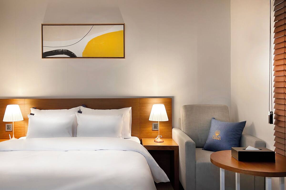 Une chambre d’hôtel avec un grand lit, deux lampes de chevet, une petite table, une chaise et une œuvre d’art accrochée au mur.