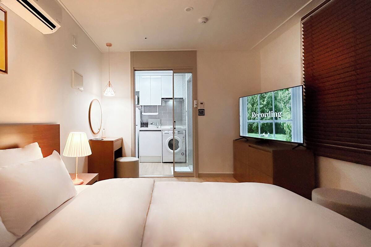 Une chambre d’hôtel équipée d’un lit, d’une télévision, d’un miroir et offrant une vue sur la cuisine et l’espace buanderie.