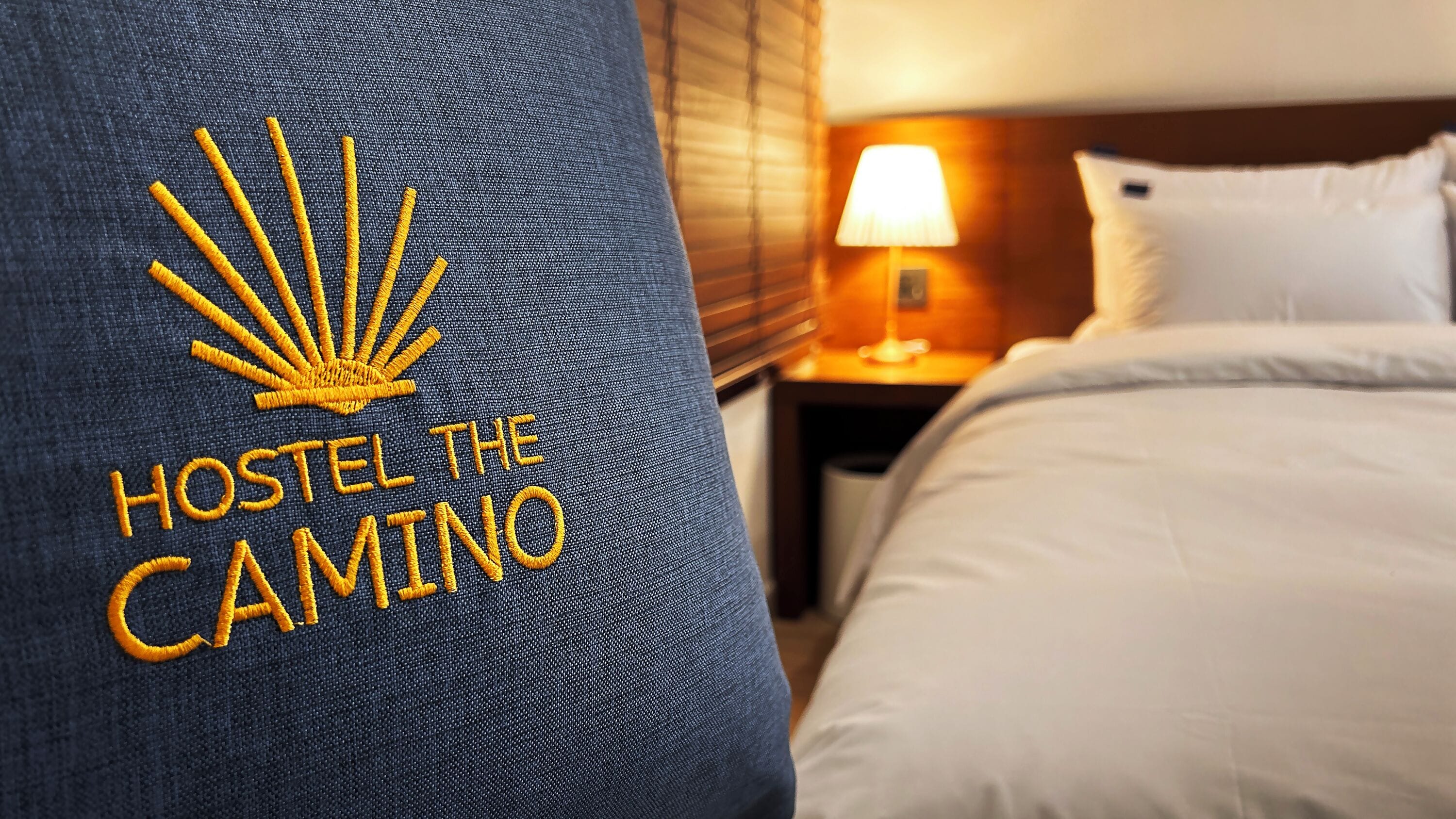 A hotel room with a bed, a nightstand, and a lamp. The pillow has embroidered text and a sun design.