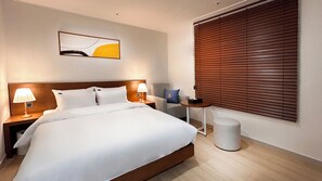 A hotel room with a large bed, two bedside lamps, a small table, a chair, a round ottoman, and a window with blinds. - Hostel The Camino Seoul (Seoul)