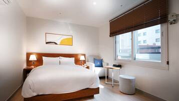 A hotel room with a large bed, bedside tables, a small table, a chair, a round stool, a window with blinds, and a painting on the wall.