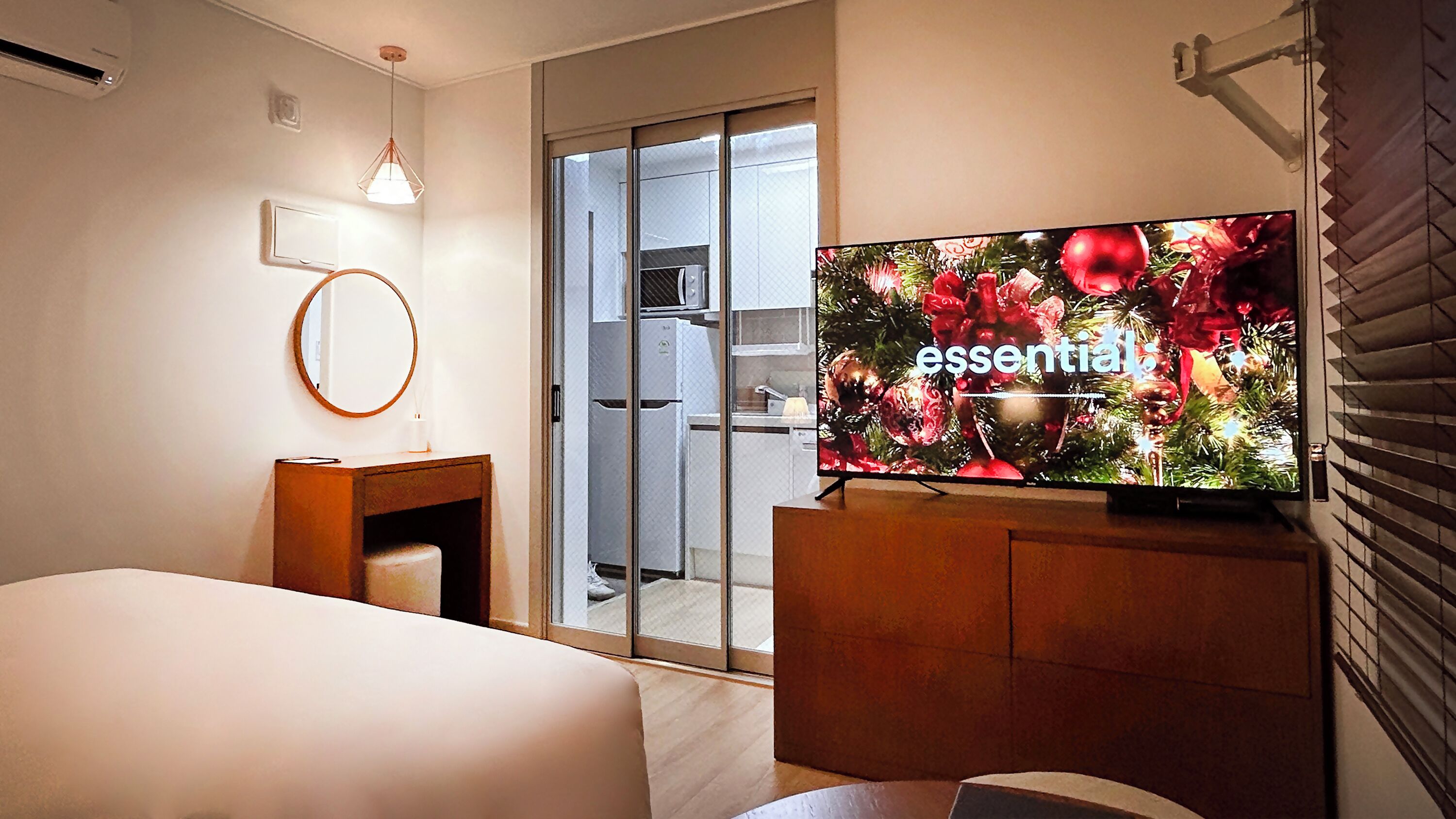 Habitación de hotel con cama, escritorio, televisión mostrando 'essential' y decoración navideña, además de una puerta corrediza que da a una zona de cocina.