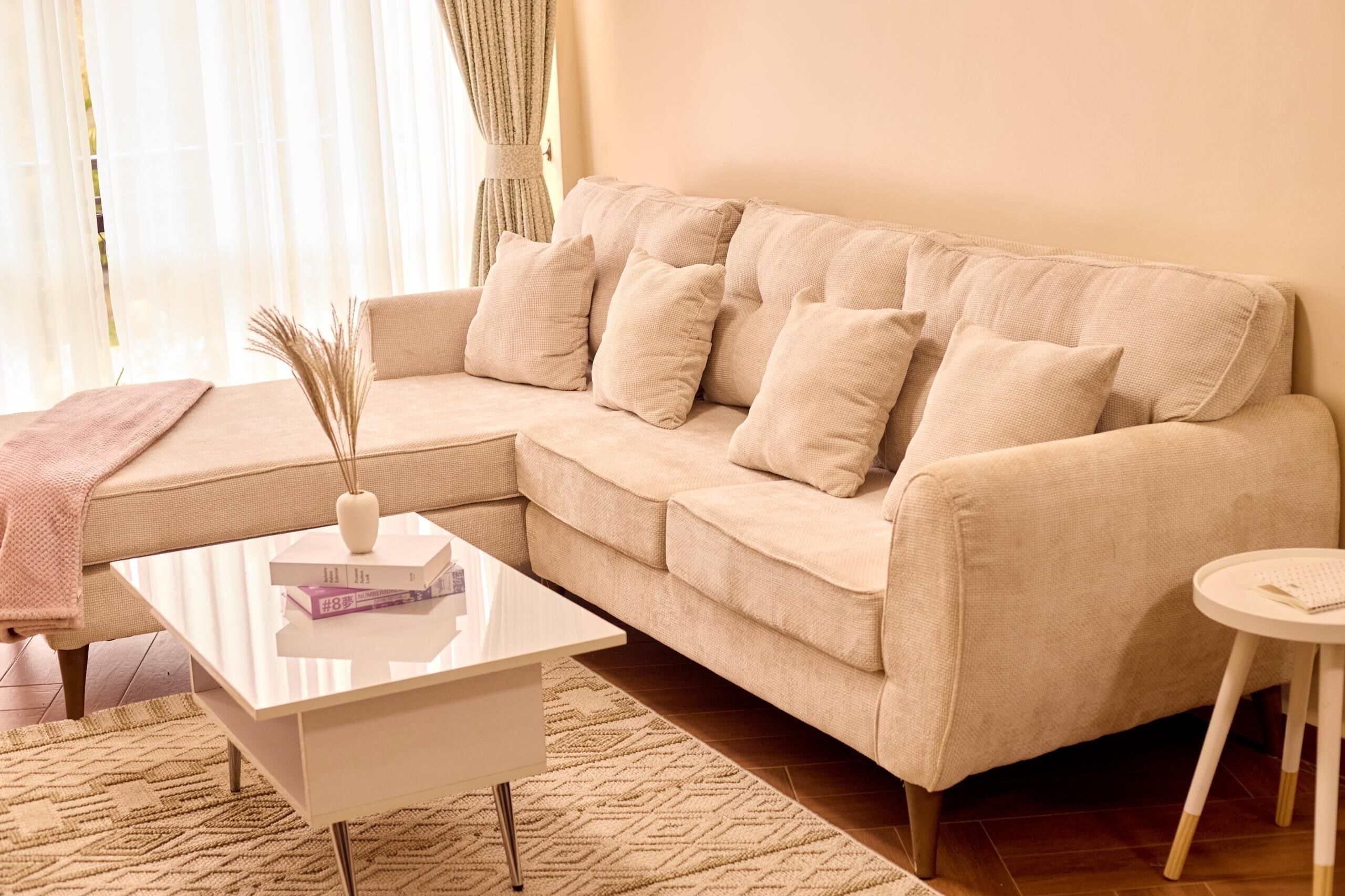 Ein beiges Sofa mit mehreren Kissen, ein weißer Couchtisch, ein kleiner runder Tisch und eine dekorative Vase mit Trockenpflanzen.