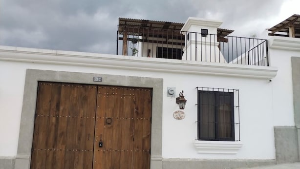 Un edificio bianco con un'ampia porta in legno a due battenti, una piccola finestra con inferriata in metallo e un balcone con ringhiera in legno.