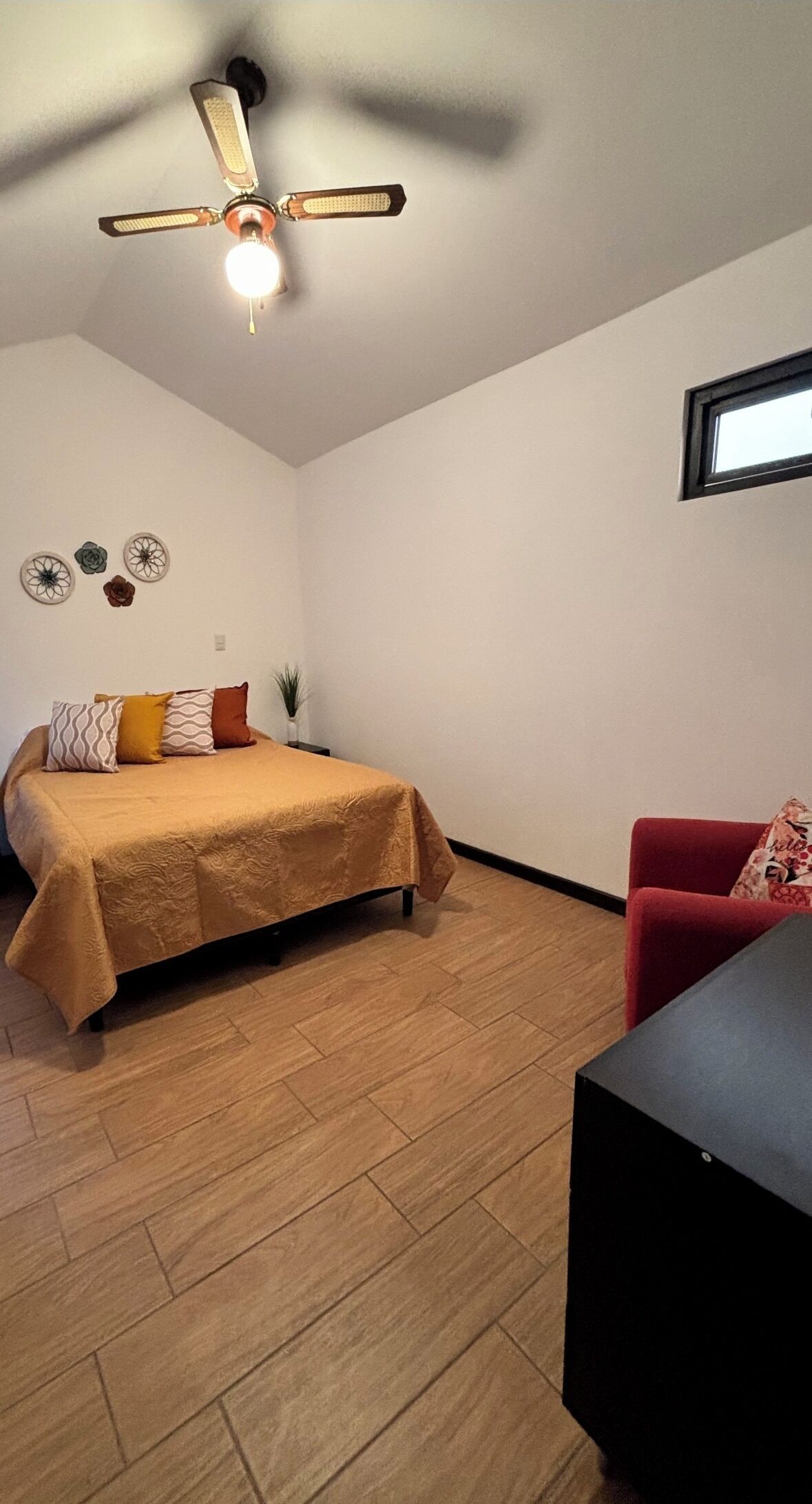 Een slaapkamer met een bed, een bureau, een rode stoel, een plafondventilator en een raam.