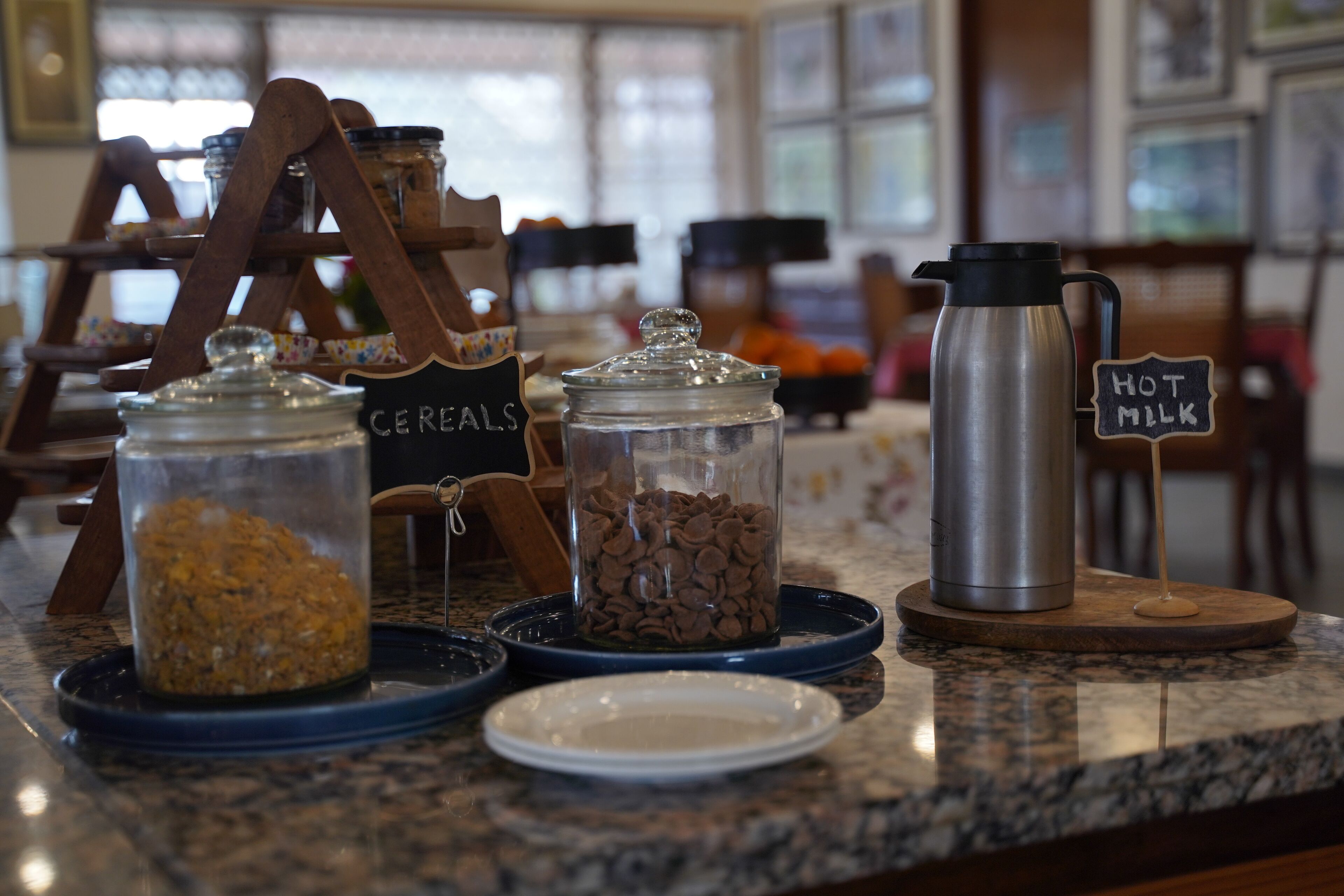A breakfast bar with labeled containers of cereals and hot milk, a coffee maker, and a wooden rack with jars.