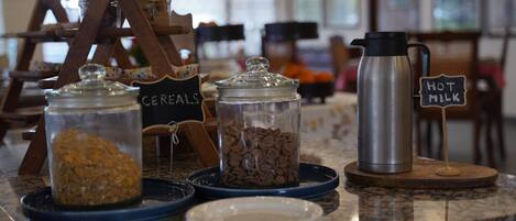 A breakfast bar with labeled containers of cereals and hot milk, a coffee maker, and a wooden rack with jars.