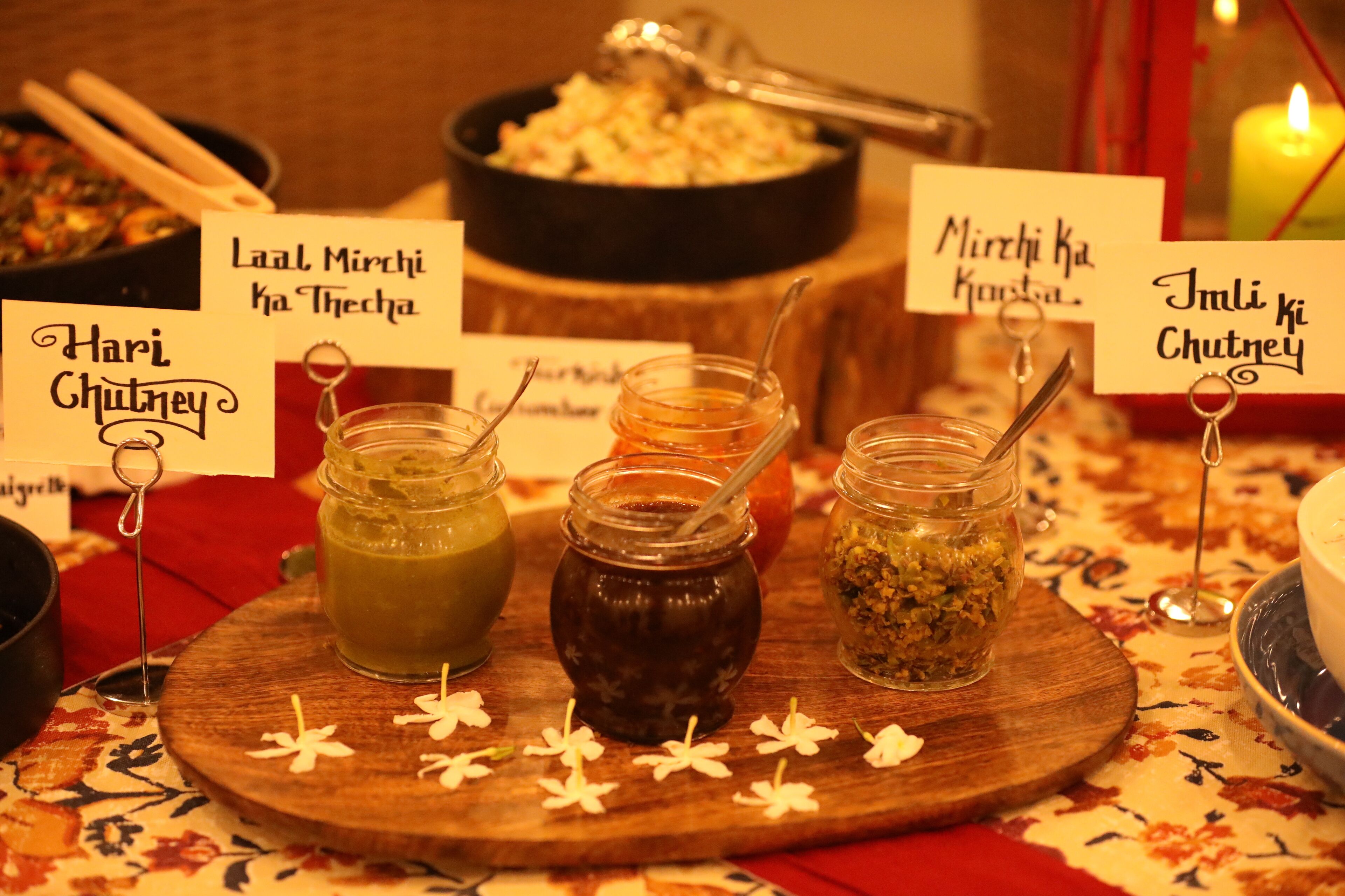 Een houten plank met drie potjes chutney, gelabeld 'Hari Chutney', 'Laal Mirchi ka Thecha' en 'Mirchi ka Chutney'.