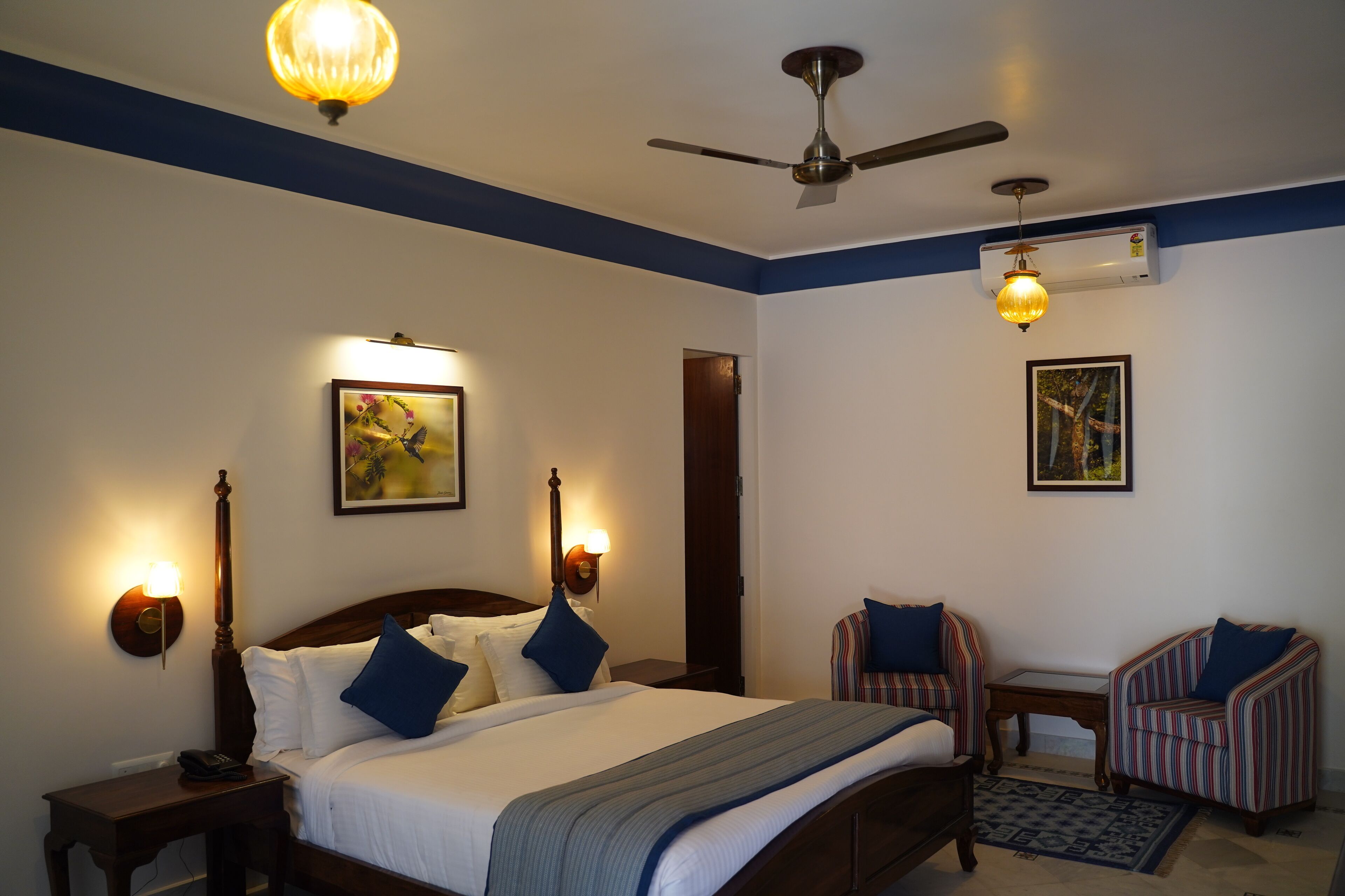 A bedroom with a bed, two chairs, a nightstand, a desk, a ceiling fan, and wall-mounted lights.