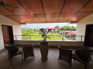 Un balcon agrémenté de fauteuils en osier et d’une plante en pot, donnant sur un jardin aménagé et un bâtiment aux toits rouges.