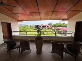 Un balcón con sillas de mimbre y una planta en maceta, con vistas a un jardín ajardinado y un edificio con tejados rojos.