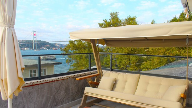 Un balcon avec un canapé et une table, donnant sur une étendue d’eau, avec un pont en arrière-plan.